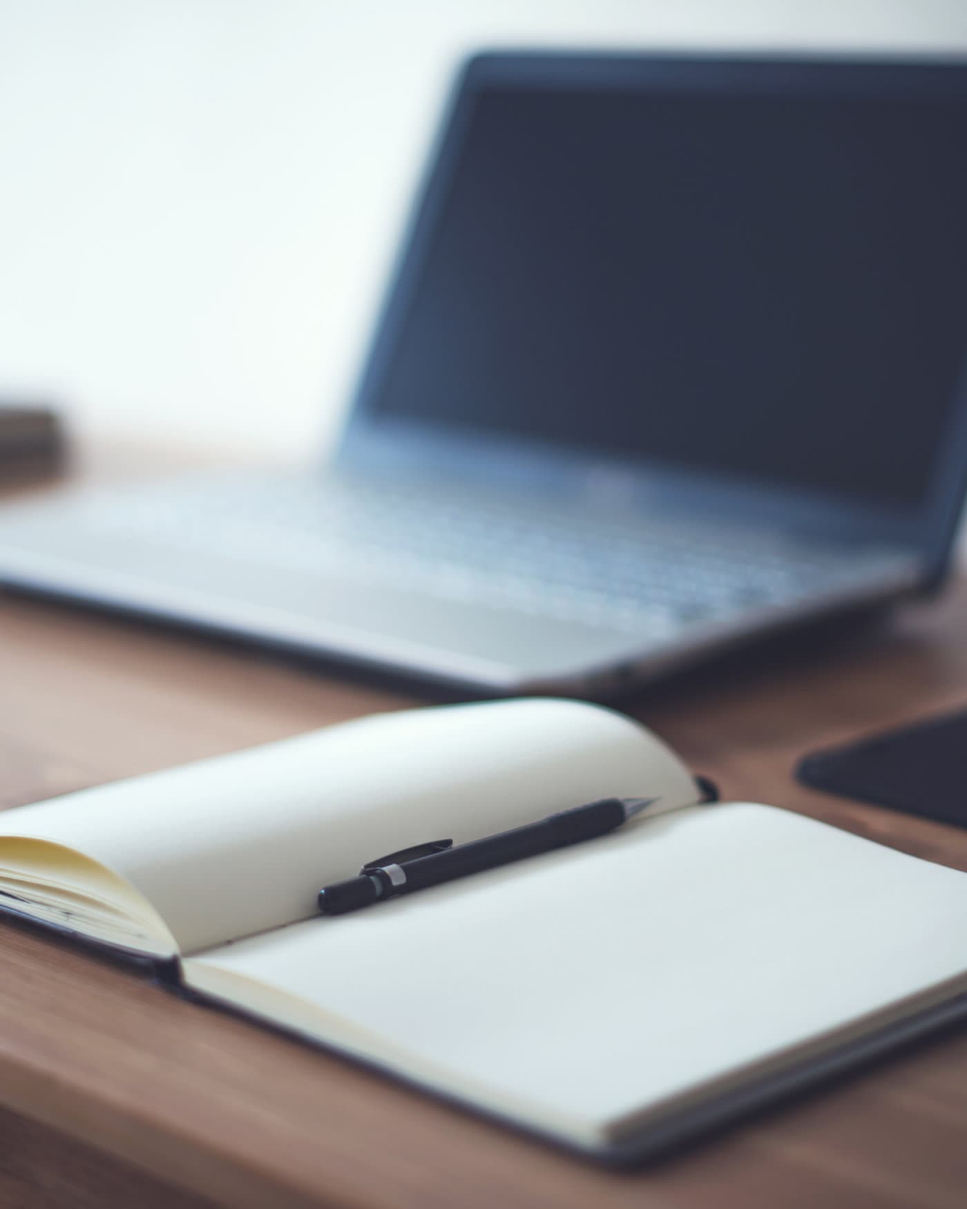 Aufgeschlagenes Notizbuch mit Stift auf einem Schreibtisch, im Hintergrund ein Laptop
